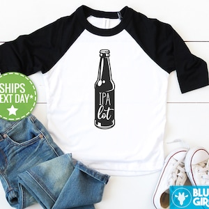 Puede incluir: Una camiseta de béisbol blanca y negra con una ilustración en blanco y negro de una botella de cerveza con el texto "IPA lot" en ella.