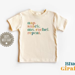 May include: A cream coloured t-shirt with the text "nap. snack. ms. rachel. repeat." in colourful letters.