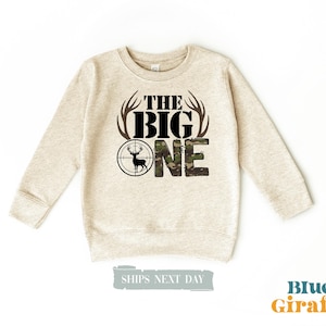 May include: A beige sweatshirt with a camouflage deer design and the text "The Big One" printed on the front.