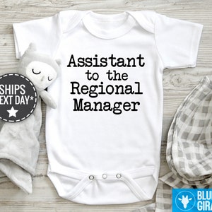 May include: White baby bodysuit with black text reading, 'Assistant to the Regional Manager'.  The bodysuit is shown on a light wood surface with a gray muslin swaddle and a gray and white gingham receiving blanket.  A small gray owl plush toy is also visible.