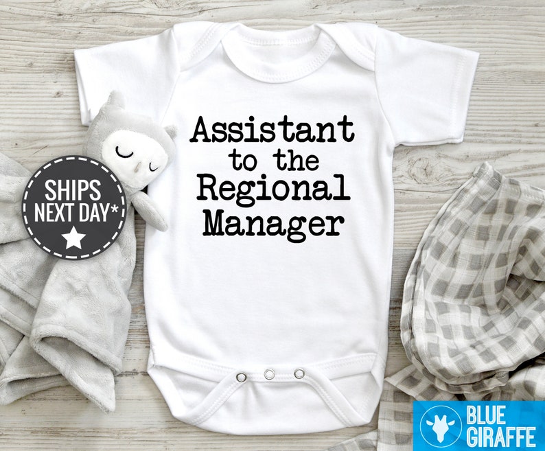 May include: White baby bodysuit with black text reading, 'Assistant to the Regional Manager'.  The bodysuit is shown on a light wood surface with a gray muslin swaddle and a gray and white gingham receiving blanket.  A small gray owl plush toy is also visible.