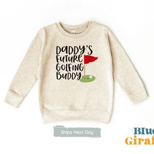 Peut inclure: Sweat-shirt beige avec le texte "Daddy's Future Golfing Buddy" en lettres noires. Un drapeau de golf rouge avec un mât blanc est représenté à côté du texte.