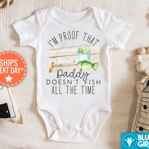 Op de afbeelding: Witte babybody met de tekst "I'M PROOF THAT Daddy DOESN'T FISH ALL THE TIME." Het ontwerp bevat een hengel, een emmer en een vis. Het Blue Giraffe-logo is zichtbaar.
