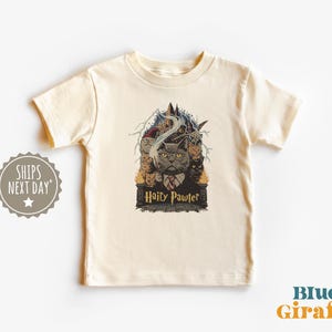 May include: Cream-colored t-shirt featuring a graphic of a cat in a suit with the text "Hairy Pawter". The design includes a castle, smoke, and other cats. The shirt is from Blue Giraffe.