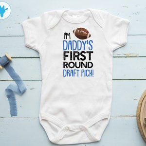 Peut inclure: Combinaison blanche pour bébé avec un texte bleu et noir qui dit "I'm Daddy's First Round Draft Pick!" avec un graphique de ballon de football brun et blanc.