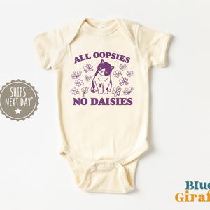 May include: A cream-colored baby onesie with a purple cat illustration and the text "All oopsies no daisies".
