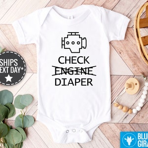 May include: White baby bodysuit featuring a black engine graphic and the text "CHECK DIAPER" with "ENGINE" crossed out. A pacifier and wooden bead clip are visible. The bodysuit is a baby clothing item.