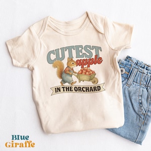 May include: A cream-colored short-sleeve bodysuit featuring a graphic of a squirrel in blue overalls pushing a wheelbarrow full of red apples. The text reads "CUTEST apple IN THE ORCHARD" in blue and red lettering, accompanied by blue denim jeans.