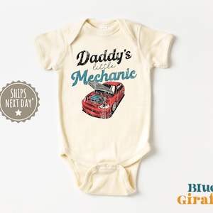 Op de afbeelding: Crèmekleurige babybody met de tekst "Daddy's little Mechanic" boven een rode autografiek. De motorkap van de auto is open en toont de motor. De tekst en afbeelding hebben een vintage uitstraling.