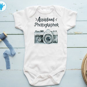 May include: White baby onesie with the text "Assistant Photographer" and a watercolor illustration of a vintage camera.