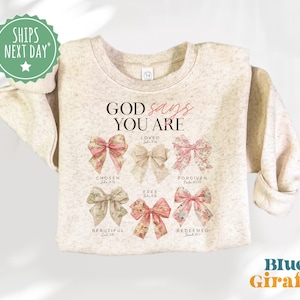May include: A beige sweatshirt with the text "GOD says YOU ARE" and a series of pink bows with bible verses underneath each bow. The bows are labeled "Loved", "Chosen", "Free", "Beautiful", "Forgiven", and "Redeemed".