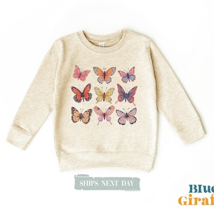 May include: A heathered beige crewneck sweatshirt featuring a graphic print of nine colorful butterflies arranged in a grid. The butterflies display various patterns and hues including orange, pink, blue, and yellow. The text Blue Giraffe is visible.