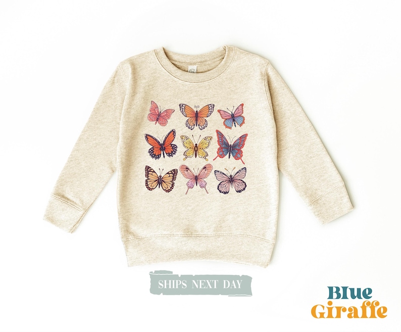 May include: A heathered beige crewneck sweatshirt featuring a graphic print of nine colorful butterflies arranged in a grid. The butterflies display various patterns and hues including orange, pink, blue, and yellow. The text Blue Giraffe is visible.