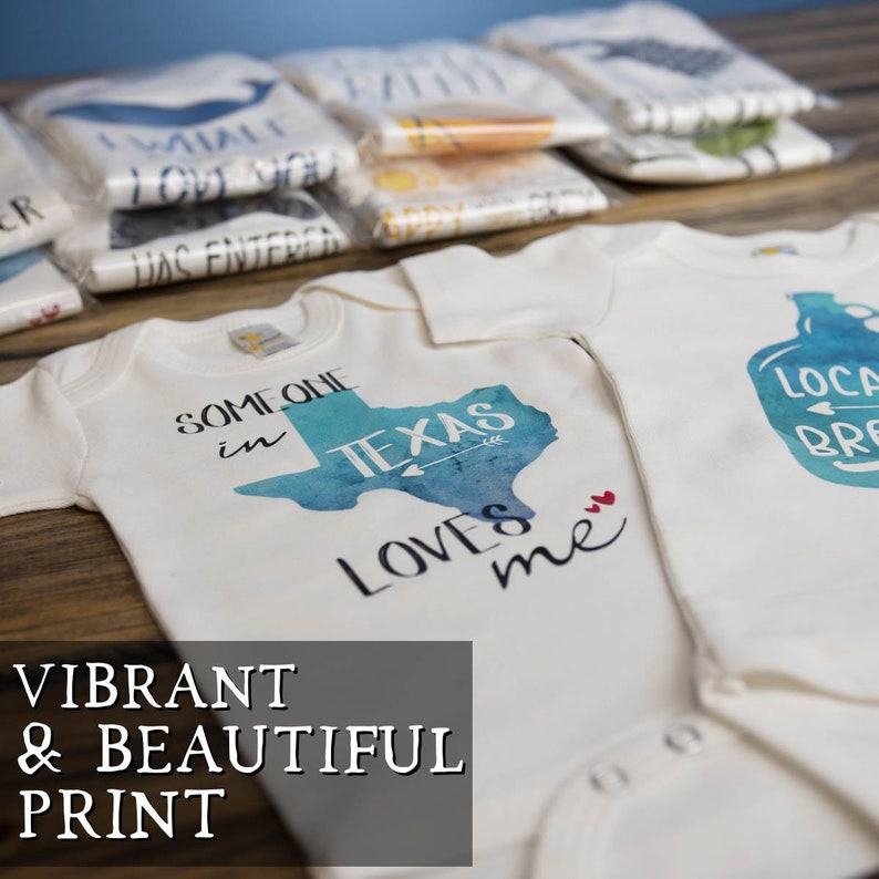 May include: Two cream-colored baby onesies feature vibrant watercolor-style prints. One onesie displays a teal Texas-shaped design with the text 'Someone in Texas Loves me.' The other onesie shows a blue mason jar graphic with the text 'Local Brew.'  Both designs are printed on soft, high-quality fabric.  The onesies are displayed on a wooden surface with stacks of folded apparel in the background, showcasing various designs and patterns.
