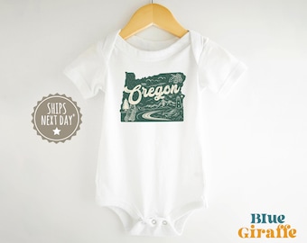 May include: White baby bodysuit with a green graphic of the state of Oregon, featuring the word "Oregon" and a scenic illustration. The bodysuit is hanging on a wooden hanger.
