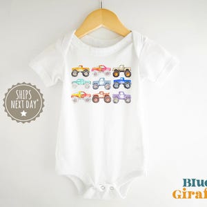 May include: White baby bodysuit with a rainbow of monster trucks printed on the front. The trucks are all different colors and have different designs. The text "Ships Next Day" is printed on the left side of the image.