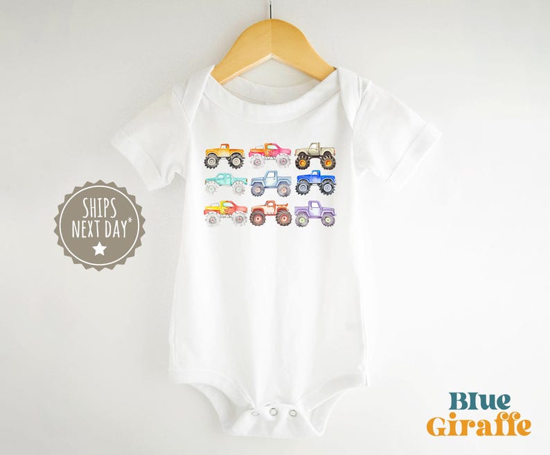 May include: White baby bodysuit with a rainbow of monster trucks printed on the front. The trucks are all different colors and have different designs. The text "Ships Next Day" is printed on the left side of the image.