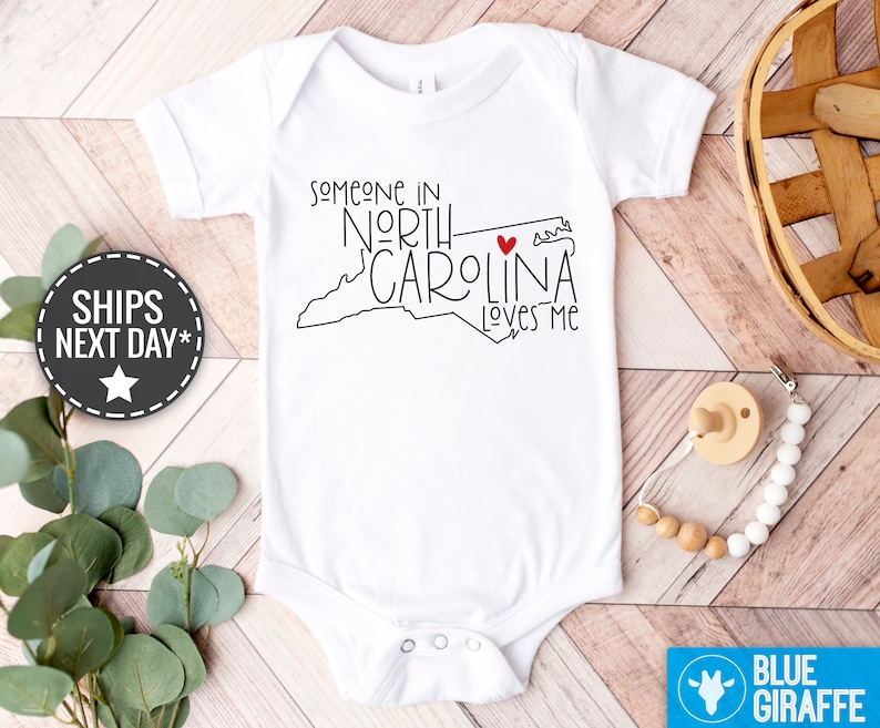 May include: White baby onesie with a black outline of the state of North Carolina and the text "Someone in North Carolina loves me" with a red heart.