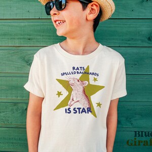 May include: Off-white t-shirt with a graphic of a rat inside a green star, with the text "RATS SPELLED BACKWARDS IS STAR". The shirt also has the text "Blue Giraffe" in the bottom right corner.