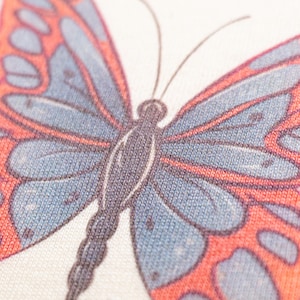 May include: A close-up view of a textile surface featuring a printed butterfly graphic. The butterfly has a dark gray body and antennae, with wings patterned in shades of muted blue and vibrant orange. The fabric texture is clearly visible throughout.