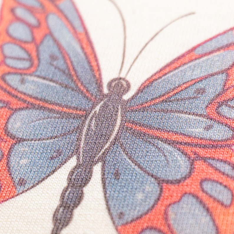 May include: A close-up view of a textile surface featuring a printed butterfly graphic. The butterfly has a dark gray body and antennae, with wings patterned in shades of muted blue and vibrant orange. The fabric texture is clearly visible throughout.