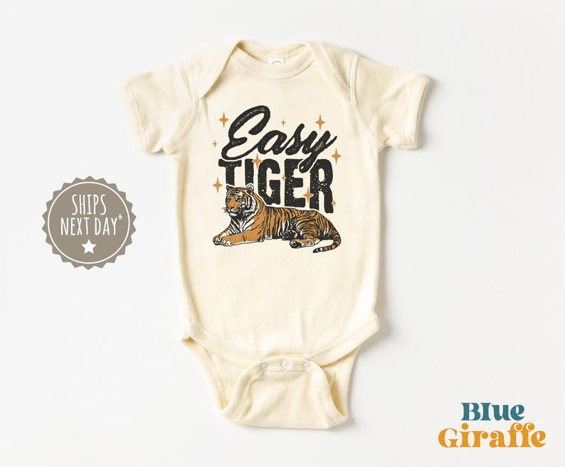 K&ouml;nnte beinhalten: Cremefarbener Baby-Body mit dem Text "Easy Tiger" in schwarzer Schrift und einer Tiger-Illustration. Der Body hat kurze &Auml;rmel und einen Druckknopfverschluss. Das Design enth&auml;lt goldene Sterne.
