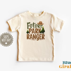May include: A cream colored t-shirt with short sleeves and the text "Future Park Ranger" in brown lettering. The text is surrounded by a green pine tree and a brown ranger hat.