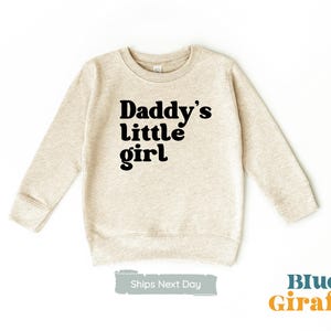 Puede incluir: Una sudadera beige con el texto "Daddy's little girl" impreso en negro.