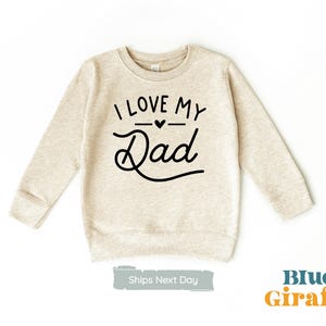 Puede incluir: Una sudadera beige con el texto "I love my Dad" en letras negras con un pequeño corazón sobre la palabra "Dad".