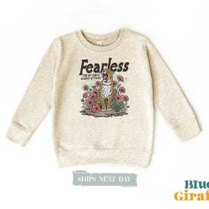 Op de afbeelding: Een lichtbeige sweatshirt met lange mouwen. De voorkant heeft een afbeelding van een luipaard omringd door rode bloemen, met de tekst "Fearless" erboven en "For my God is always with me" eronder.