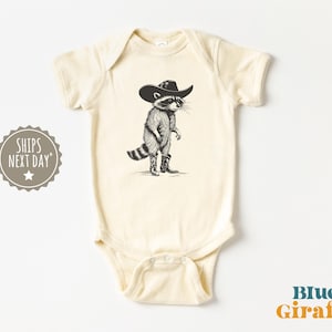 May include: A cream-colored short-sleeve baby bodysuit featuring a black and white graphic illustration of a raccoon wearing a cowboy hat and cowboy boots. The design is centered on the chest of the garment, which has a round neckline and snap closures.
