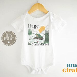 Puede incluir: Un body blanco de manga corta para beb&eacute; con una ilustraci&oacute;n gr&aacute;fica de una rana verde sentada sobre una roca en un estanque con juncos y un sol amarillo. El texto sobre el gr&aacute;fico dice Rage y el texto debajo del gr&aacute;fico dice Consumes Me.