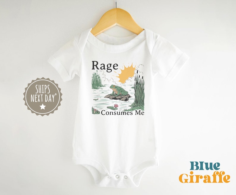 Puede incluir: Un body blanco de manga corta para beb&eacute; con una ilustraci&oacute;n gr&aacute;fica de una rana verde sentada sobre una roca en un estanque con juncos y un sol amarillo. El texto sobre el gr&aacute;fico dice Rage y el texto debajo del gr&aacute;fico dice Consumes Me.