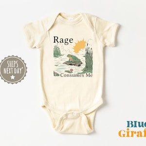 Puede incluir: Un body de beb&eacute; de manga corta color crema con una ilustraci&oacute;n gr&aacute;fica de una rana verde sentada sobre una roca en un estanque con juncos y un sol amarillo. El texto "Rage Consumes Me" est&aacute; impreso en negro sobre y bajo la ilustraci&oacute;n.