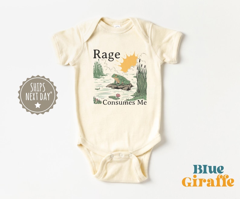 Puede incluir: Un body de beb&eacute; de manga corta color crema con una ilustraci&oacute;n gr&aacute;fica de una rana verde sentada sobre una roca en un estanque con juncos y un sol amarillo. El texto "Rage Consumes Me" est&aacute; impreso en negro sobre y bajo la ilustraci&oacute;n.