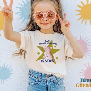 May include: A cream-colored t-shirt with the text "RATS SPELLED BACKWARDS IS STAR" and a graphic of a rat inside a green star. The shirt is paired with blue jeans. The background features colorful sunburst designs.