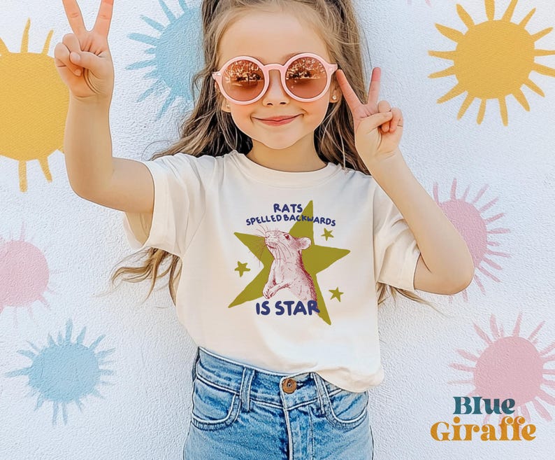 May include: A cream-colored t-shirt with the text "RATS SPELLED BACKWARDS IS STAR" and a graphic of a rat inside a green star. The shirt is paired with blue jeans. The background features colorful sunburst designs.