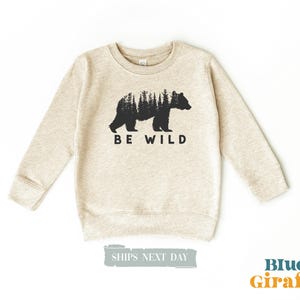 May include: A beige sweatshirt with a black bear silhouette and the text "BE WILD" printed on the front.