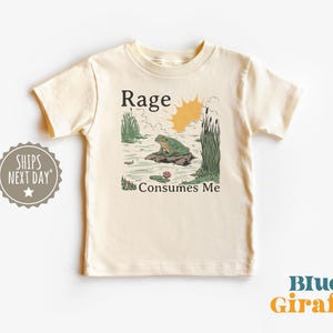 Puede incluir: Una camiseta de manga corta color crema con una ilustraci&oacute;n gr&aacute;fica de una rana verde sentada sobre una roca en un estanque con juncos y un sol amarillo. El texto "Rage" est&aacute; impreso sobre la rana y "Consumes Me" debajo de la rana.