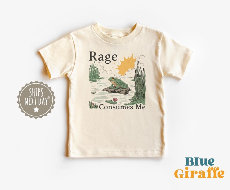 Puede incluir: Una camiseta de manga corta color crema con una ilustraci&oacute;n gr&aacute;fica de una rana verde sentada sobre una roca en un estanque con juncos y un sol amarillo. El texto "Rage" est&aacute; impreso sobre la rana y "Consumes Me" debajo de la rana.