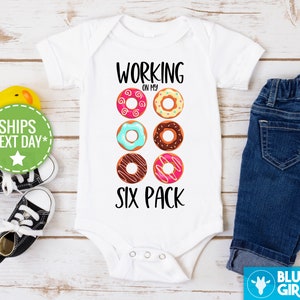 May include: White baby onesie with the text "Working on my six pack" and a graphic of six colorful donuts.