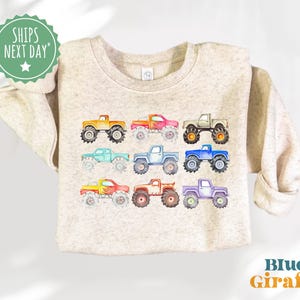 May include: A beige sweatshirt with a colorful design of monster trucks. The trucks are in various colors, including red, orange, yellow, green, blue, and purple. The sweatshirt has a crew neck and long sleeves.