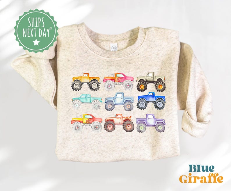 May include: A beige sweatshirt with a colorful design of monster trucks. The trucks are in various colors, including red, orange, yellow, green, blue, and purple. The sweatshirt has a crew neck and long sleeves.