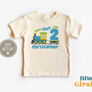 May include: A cream-colored toddler t-shirt with a colorful train graphic. The design includes the text "choo choo I'm 2" and the name "Christopher" in blue. The train is blue, yellow, and green.