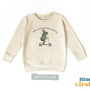 May include: Beige crewneck sweatshirt with a graphic of a green frog wearing a wizard hat and riding a scooter. The text "Off to commit tom foolery" is above the frog. The Blue Giraffe logo is in the bottom right.