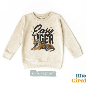K&ouml;nnte beinhalten: Ein hellbeige Sweatshirt mit langen &Auml;rmeln. Auf der Vorderseite steht der Text "Easy Tiger" in Schwarz mit einer Tiger-Illustration. Der Tiger ist orange und schwarz. Das Sweatshirt ist ein Kinderbekleidungsartikel.