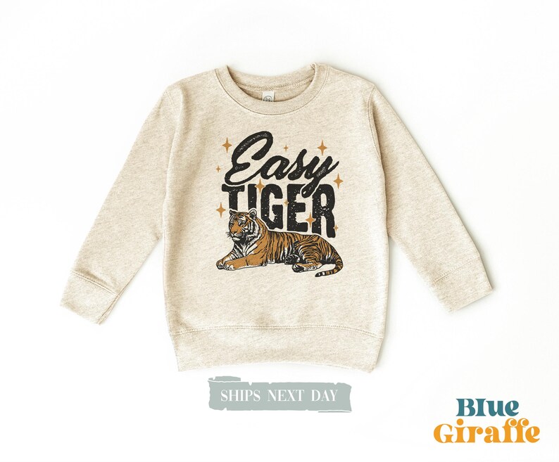 K&ouml;nnte beinhalten: Ein hellbeige Sweatshirt mit langen &Auml;rmeln. Auf der Vorderseite steht der Text "Easy Tiger" in Schwarz mit einer Tiger-Illustration. Der Tiger ist orange und schwarz. Das Sweatshirt ist ein Kinderbekleidungsartikel.