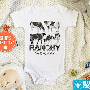 May include: White baby onesie with a black and white graphic of cows and the text "Ranchy Stuff".