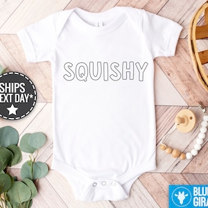 Puede incluir: Un body blanco para bebé con el contorno negro de la palabra "SQUISHY" impreso en la parte delantera.