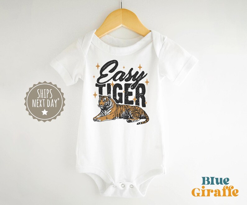 K&ouml;nnte beinhalten: Wei&szlig;er Baby-Body mit dem Text "Easy Tiger" in schwarzer und goldener Schrift, mit einer Tiger-Grafik. Der Body h&auml;ngt an einem Holzb&uuml;gel.